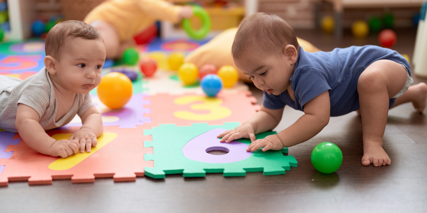 Two young infants play together on a foam mat with number cutouts and colourful balls