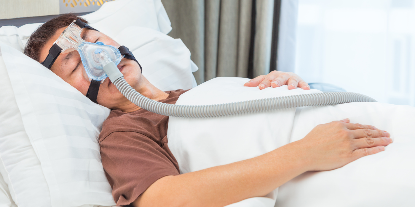 A man in a brown shirt, sleeping on a white pillow, with white covers wears a CPAP mask.