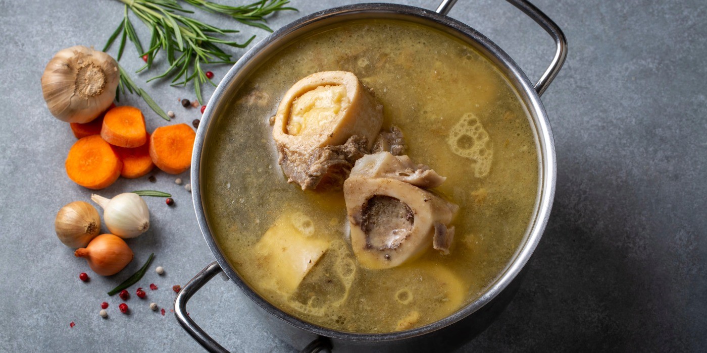 A steel pot of golden liquid, bones and other ingredients on a counter with some garlic, onion, carrots and rosemary.