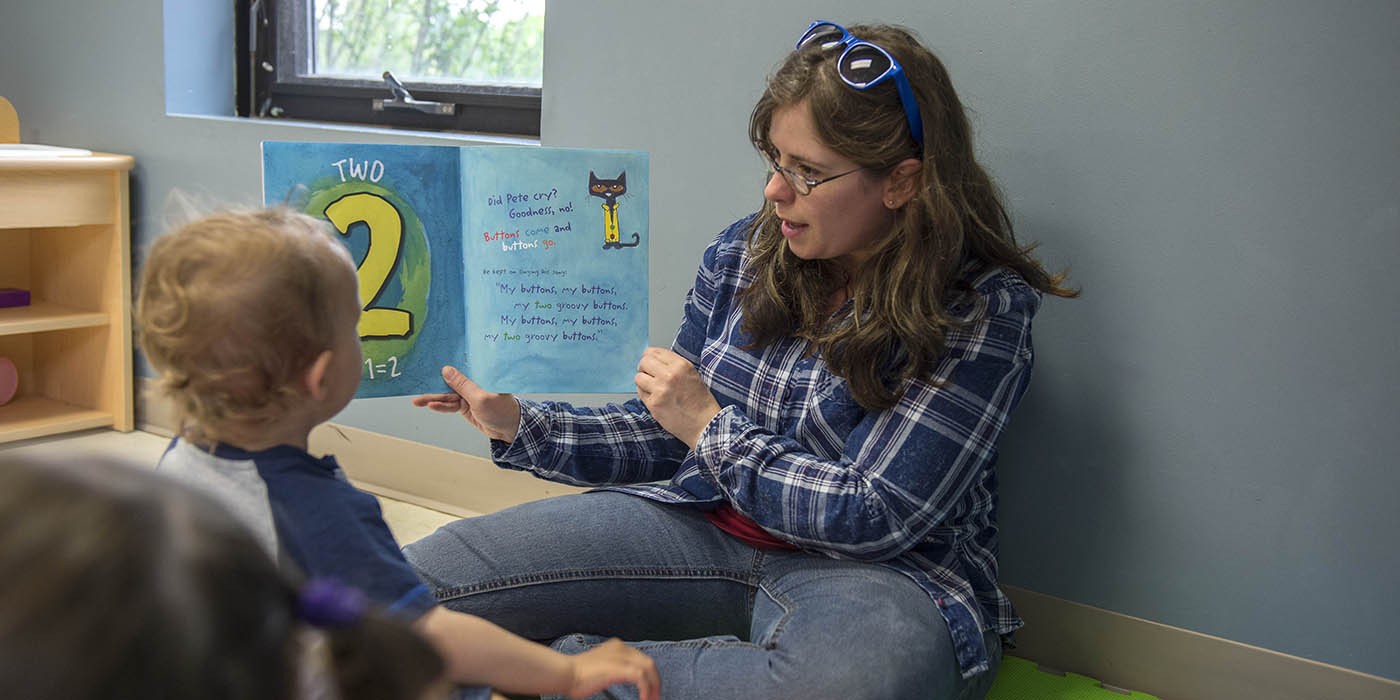 An individual sits on the floor of a classroom at the Simcoe/Norfolk Regional Campus, reading a book to a small group of children as part of the Early Childhood Education program; the book is open to a page with the number "2" and a cartoon cat.