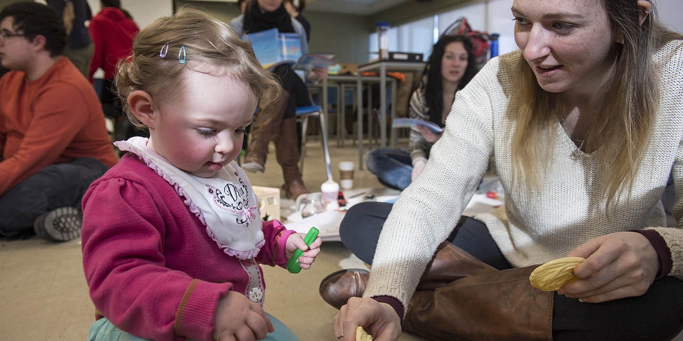 Inside a classroom at Fanshawe College's School of Community Studies, a child in a pink sweater and turquoise pants sits on the floor, holding a green toy. A person in a cream-colored sweater and brown boots sits cross-legged nearby, holding a yellow toy. Other people are seated in the background.