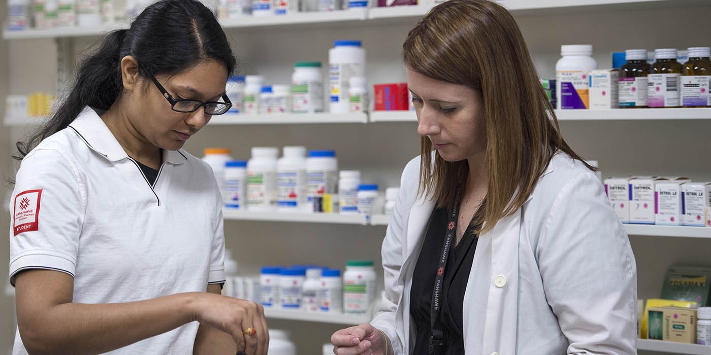 At the School of Health Sciences of Fanshawe College at the London Campus, two people stand in front of shelves stocked with various medications; one person holds a small container, while the other holds a few pills.
