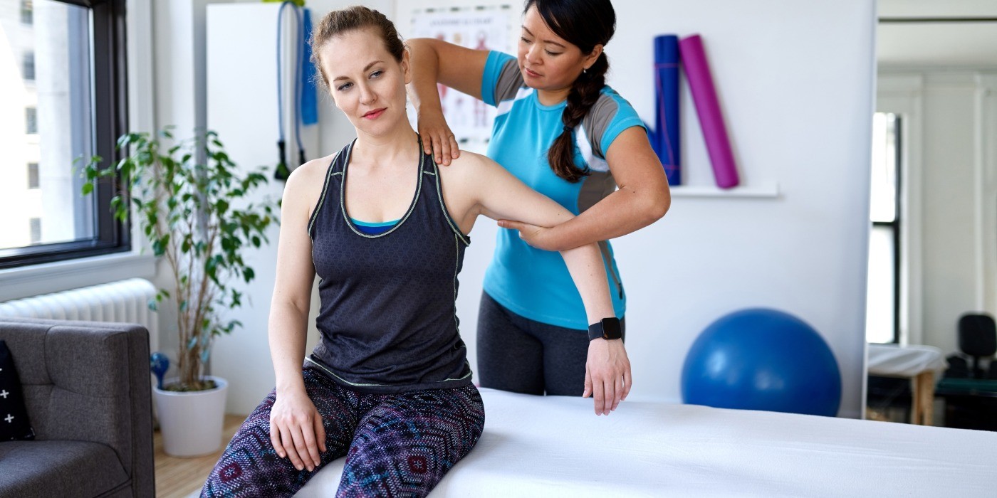 A woman sits on a therapy table, as a physiotherapy practitioner works on her shoulder.  A blue yoga ball is blurred in the background along with an indoor plant, yoga mats and tension cords.