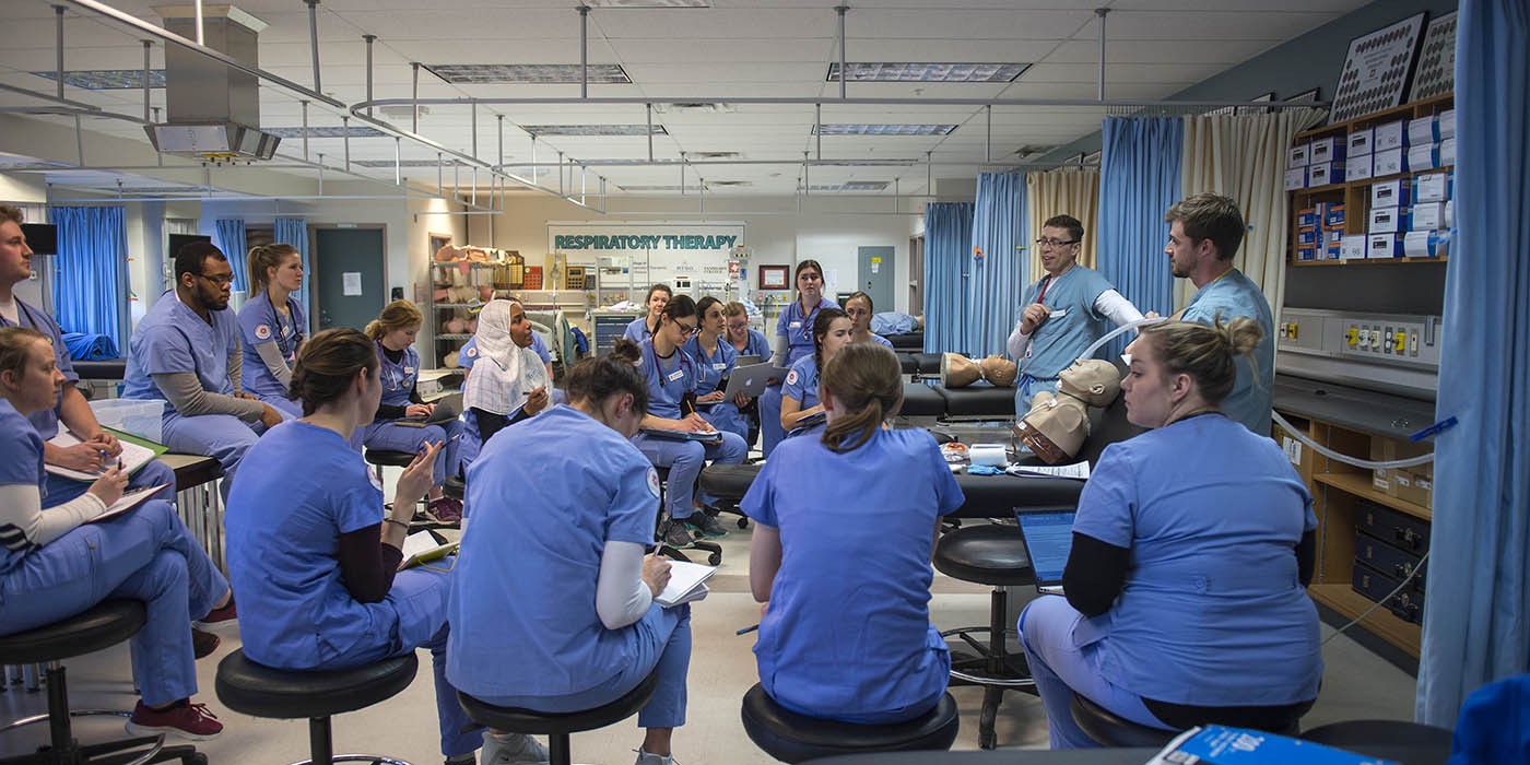 In a classroom at the School of Health Sciences of Fanshawe College at the London Campus, a group of people in blue scrubs sit on rolling stools, facing two people who are demonstrating a procedure on a