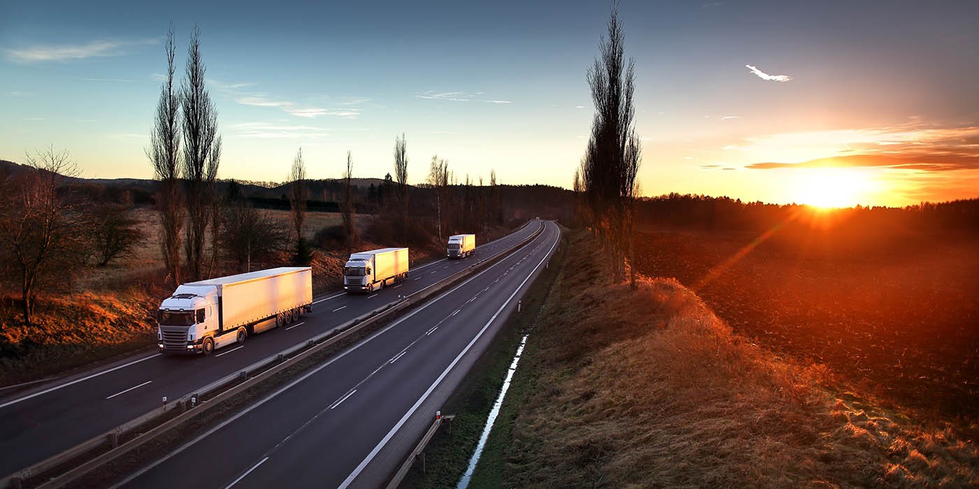 The image shows a multi-lane highway with three large trucks driving on it, with a field and trees on either side, and a bright sunset in the distance.