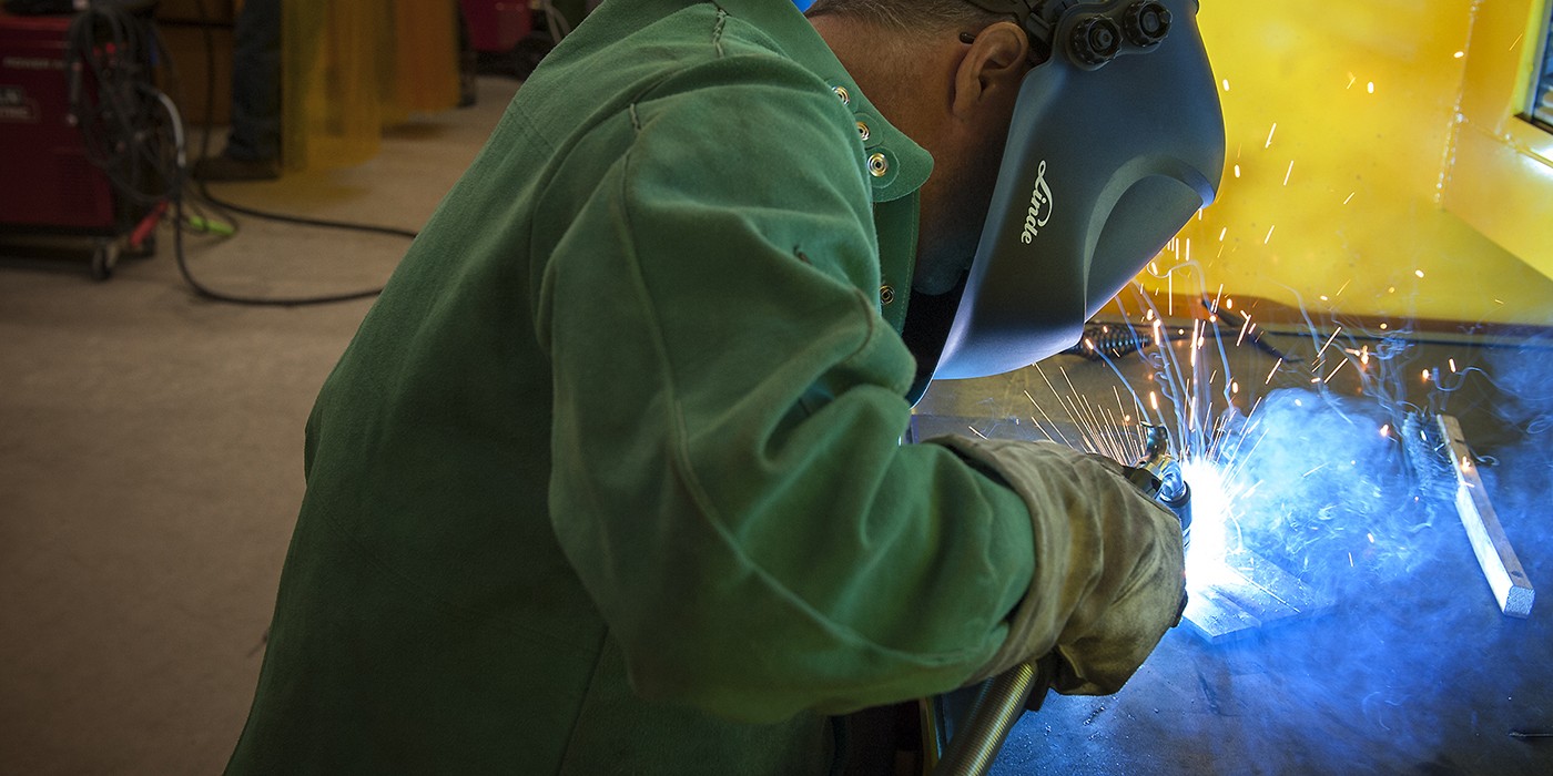 A person wearing a green jacket, gloves, and a welding mask is welding metal, creating bright sparks and blue light. The person is positioned in front of a yellow wall, with welding equipment and gas tanks visible in the background.