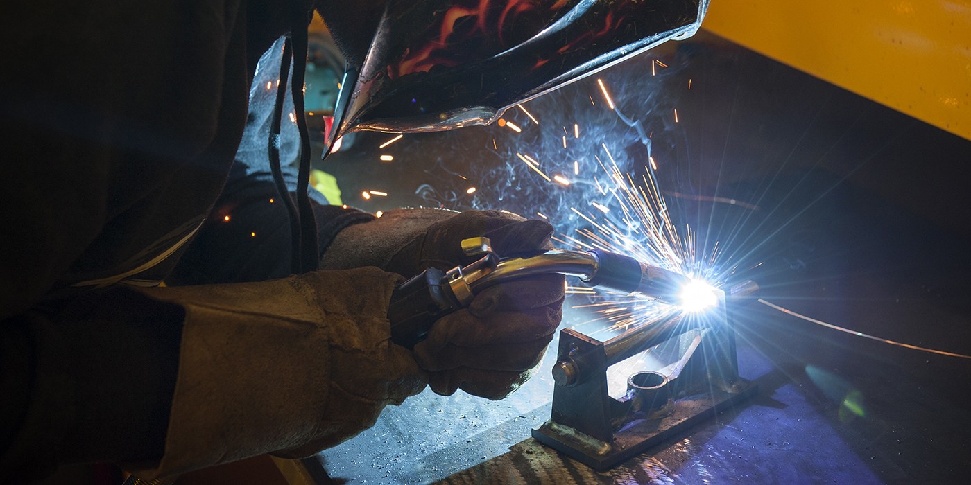 A person wearing a flame-patterned welding mask and protective gloves welds metal, creating bright sparks and smoke.