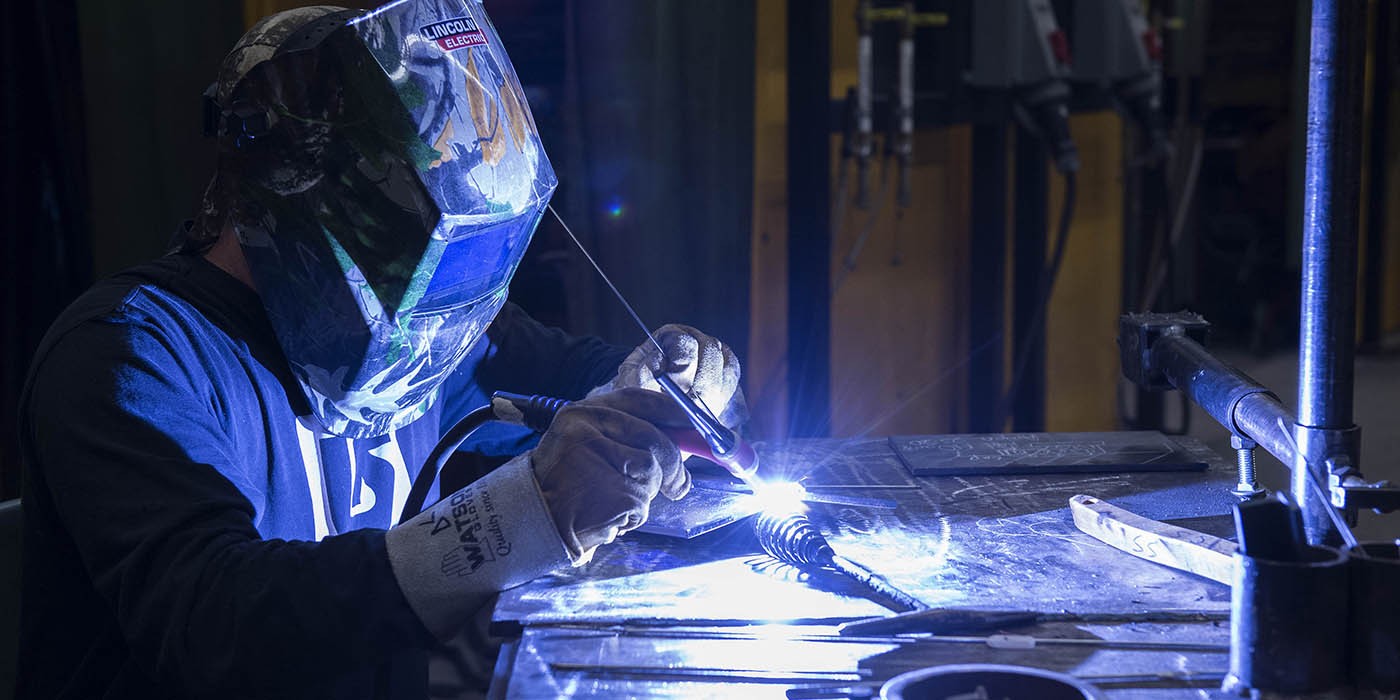A person wearing a welding mask and gloves is welding metal on a table, with bright sparks flying.
