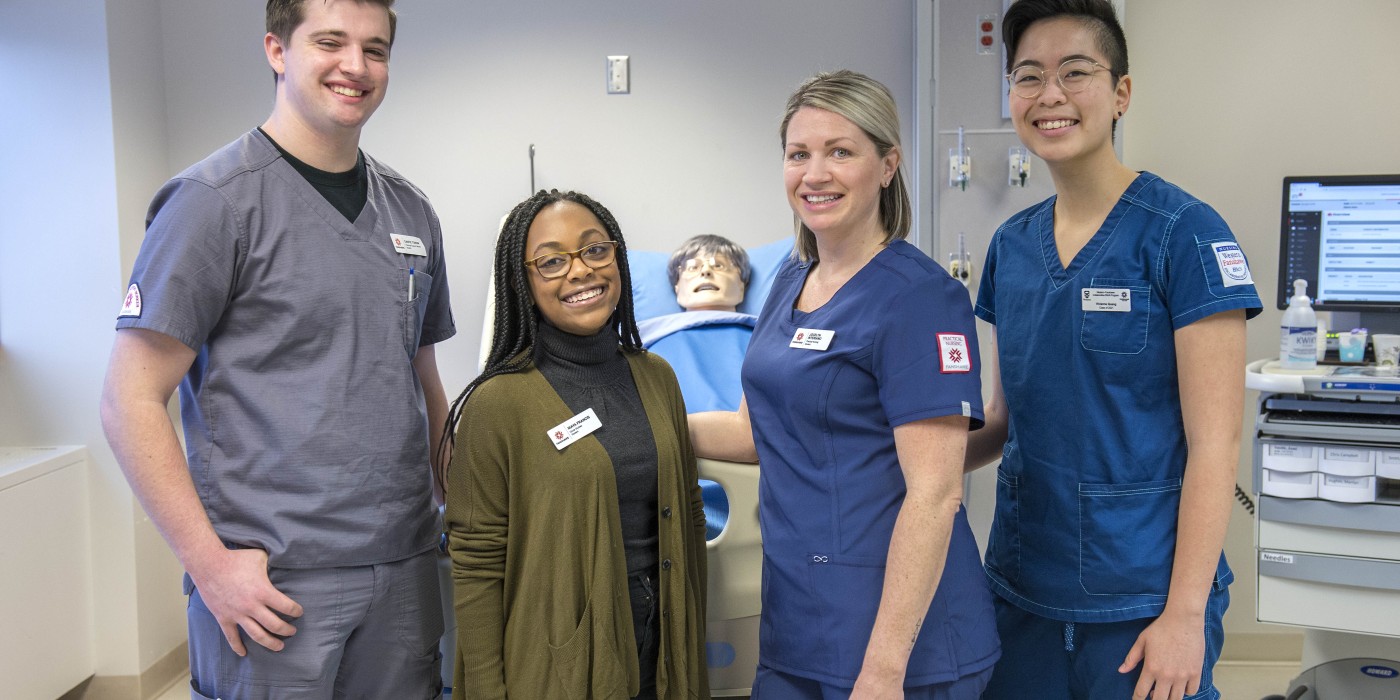 In a simulated hospital room at Fanshawe College's School of Nursing at the London Campus, four people stand smiling in front of a hospital bed with a mannequin lying in it, with medical equipment visible in the background.