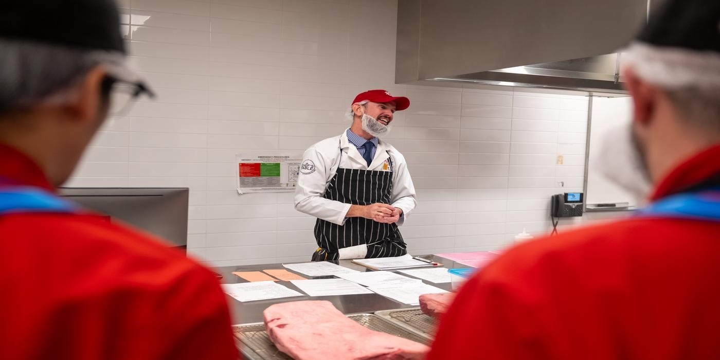 At the London Downtown Campus, in a room with white tiled walls and stainless steel counters, a person in a white lab coat, red baseball cap, and black and white striped apron stands behind a counter with papers and a large cut of meat, while two people in red jackets stand in the foreground.