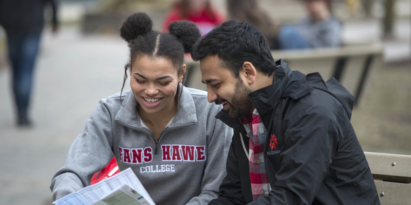 Two people wearing Fanshawe College branded clothing look at a map while sitting on a bench at Fanshawe College's London Campus.