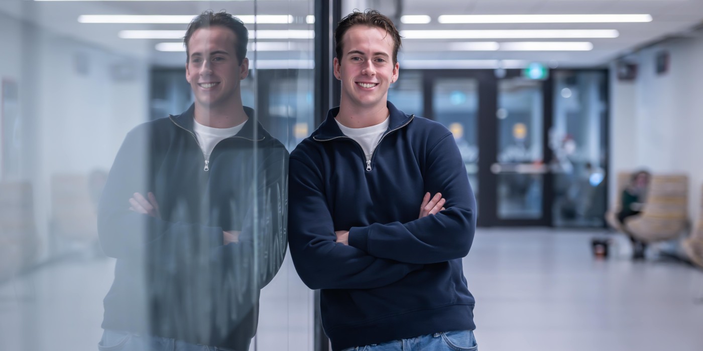 A person in a navy sweater and jeans stands with arms crossed, leaning against a glass wall that reflects their image.