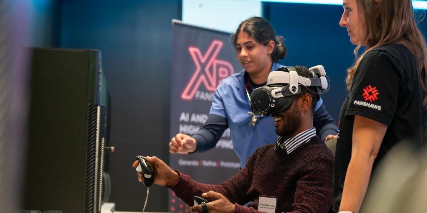 A person wearing a virtual reality headset and holding handheld controllers sits at a desk while two others observe nearby.