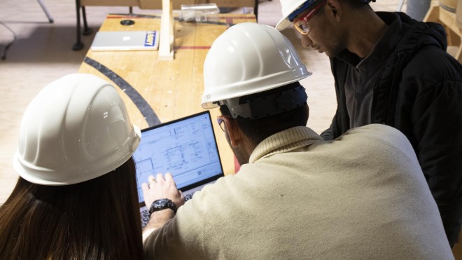 Two students with Construction Project Management instructor, reviewing plans on laptop at job site