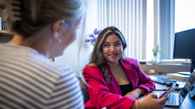 Woman sitting at desk with cell phone, conversing with another person.