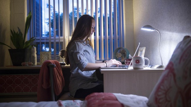 A student sitting at a desk in their dorm room working on a laptop