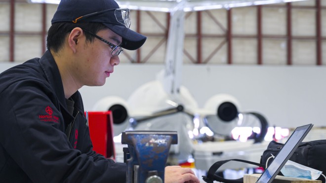 Aviation student sitting at a laptop with a plane in the background.