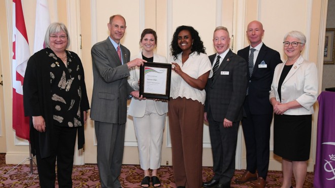 A group of seven people, including His Royal Highness Prince Edward The Duke of Edinburgh, are lined up smiling at the camera next to a Canadian flag.