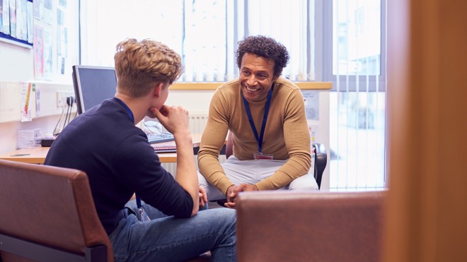 A counselor and a young man sit in an office talking. The counselor smiles warmly, creating a supportive and empathetic atmosphere.