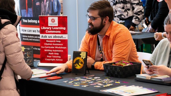 A man in an orange shirt sits at a table covered with colorful pins and brochures. A red sign reads "Library Learning Commons." The atmosphere is interactive.