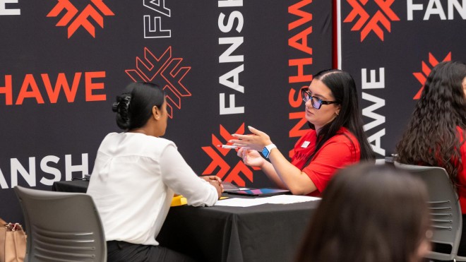 Two individuals sit at a table with a black tablecloth, in front of a backdrop with a repeating pattern