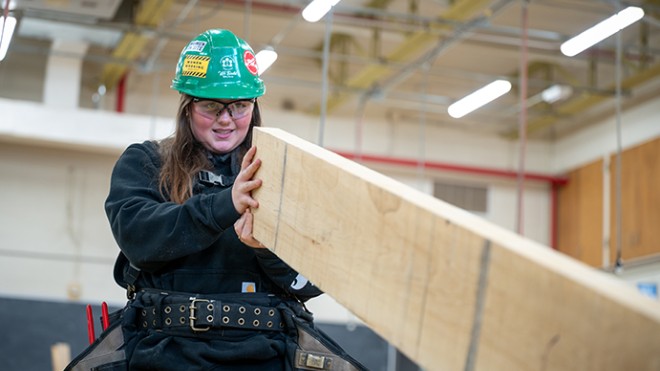 A female tradesperson assesses a piece of lumber in a shop environment.