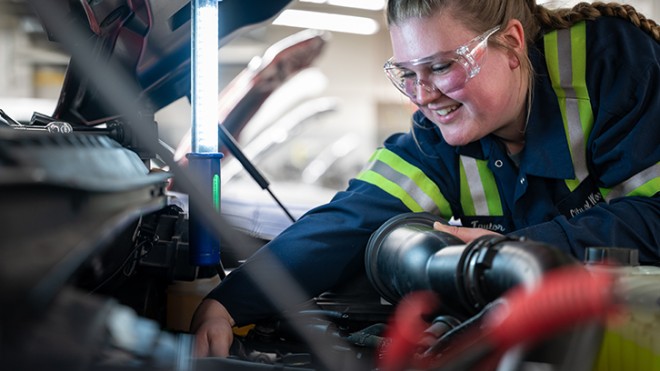 A woman working on the engine of a vehicle, under a propped up hood.