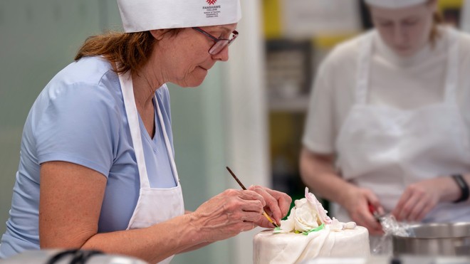 Two students in aprons carefully paint food colouring onto iced cakes