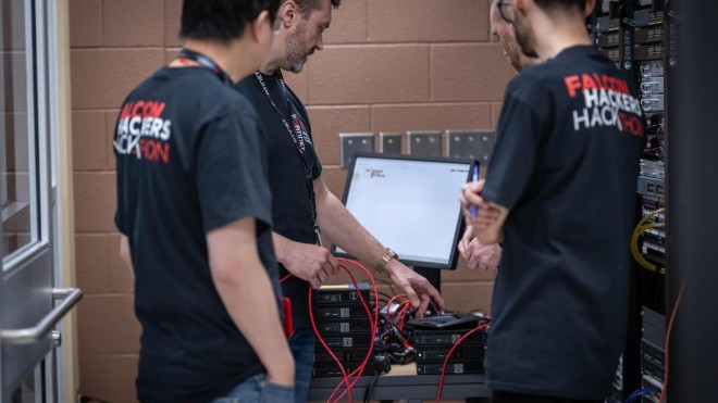 At the London Campus of Fanshawe College, three individuals wearing matching shirts stand around a table with a computer monitor and several small computers connected with red cables, next to a rack of network equipment.