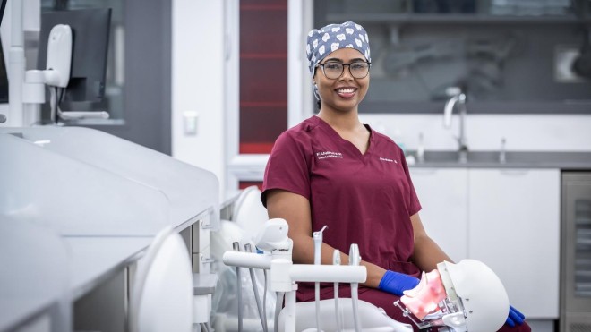 In the School of Health Sciences at Fanshawe College's London Campus, someone wearing a burgundy scrub top and a surgical cap patterned with teeth sits in a dental chair, with a dental mannequin in their lap.