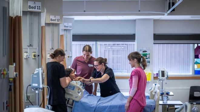 In a bright hospital room, four individuals in scrubs surround a patient in a bed with blue linens, one using a stethoscope