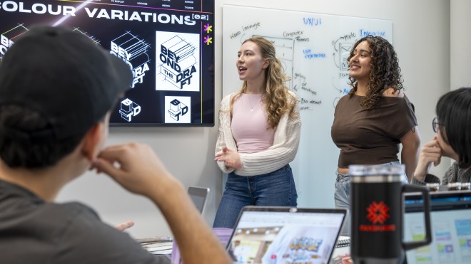 A group of individuals is gathered in a room, facing a large screen displaying "COLOUR VARIATIONS" and graphic designs, including a "BEYOND THE DRAFT" logo. To the right, a whiteboard is covered with UX/UI notes and diagrams. Two people stand near the screens, one speaking with hands gesturing, the other smiling. In the foreground, the back of a person's head and shoulder are visible, along with open laptops and a black travel mug on a table.