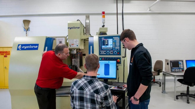 Inside a Fanshawe College lab, three people are gathered around a Cyclone machine and a computer station, reviewing a design on the screen.
