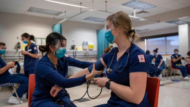 Inside a classroom at Fanshawe College's London Campus School of Nursing, two people wearing blue scrubs, face masks, and face shields are seated on orange chairs, practicing taking blood pressure. The person on the right has a stethoscope around their neck and is holding the blood pressure cuff, while the person on the left has their arm extended for the reading. Other people in scrubs are seated in the background, also practicing.