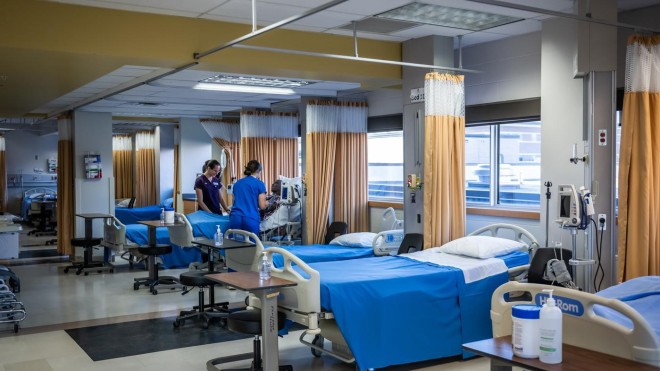A hospital ward features multiple beds with blue linens, separated by privacy curtains. Medical equipment and small tables are positioned near the beds. Overhead lighting illuminates the room.