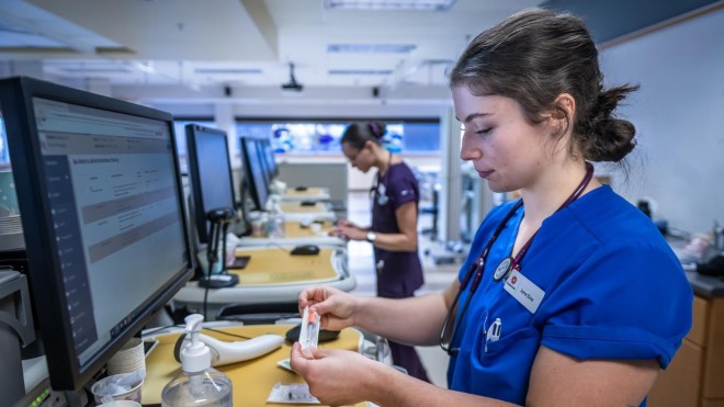A person in blue scrubs holds a syringe, standing at a desk with a computer monitor, hand sanitizer, and medical supplies. Another person in purple scrubs works at a computer in the background.