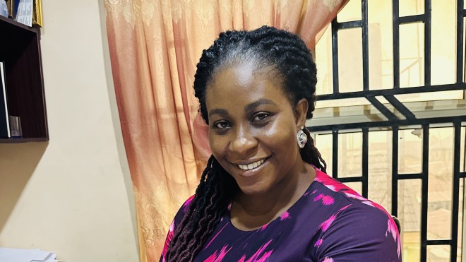 A woman in a pink and purple top smiles as she sits behind a desk full of papers.