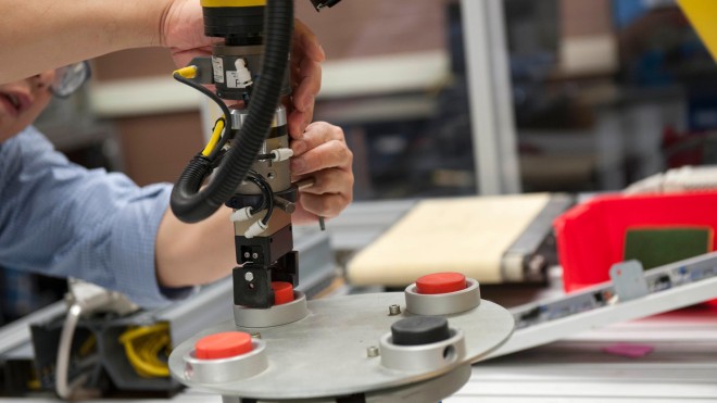 A robotic arm is being adjusted above a circular platform with red and black buttons, with a conveyor belt visible in the background.