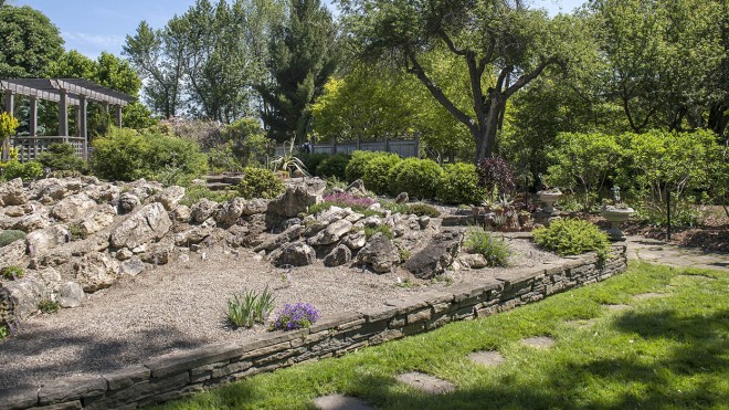 The Mac Cuddy Botanic Garden features a rock garden with a gravel bed, surrounded by a low stone wall and lush greenery, including a wooden pergola in the background.