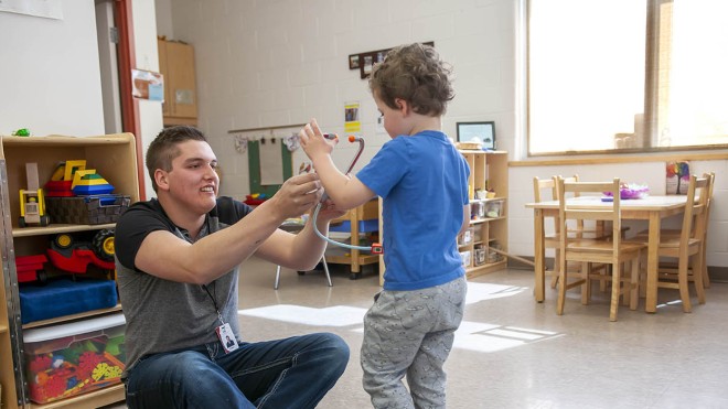 At the Huron-Bruce Regional Site of Fanshawe College in Clinton, someone sits on a rug in the Early Childhood Education program classroom, holding a toy stethoscope to a child's chest. A toy tool box sits on the rug, and shelves of toys are in the background. A small table and chairs sit near a window.
