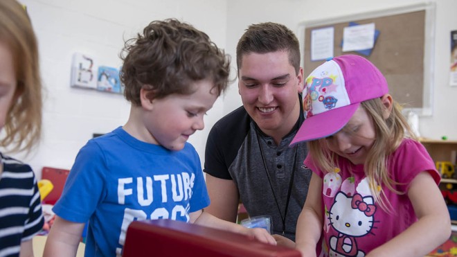 Inside a classroom at the Huron-Bruce Regional Site of Fanshawe College in Clinton, an Early Childhood Education program event shows a smiling person with short brown hair, wearing a lanyard, leaning over a red toy with two children. One child wears a blue shirt with the word "FUTURE" on it, and the other child wears a pink shirt and a pink baseball cap. A clock hangs on the wall in the background.
