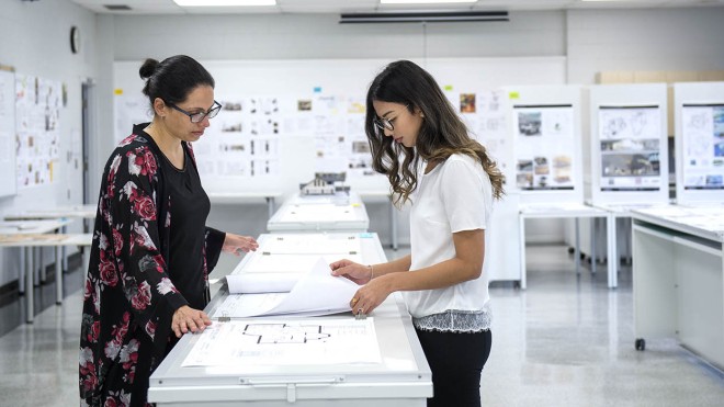 Two people stand at a table reviewing architectural drawings in a bright classroom at the London Campus of Fanshawe College. Whiteboards and presentation boards line the walls.