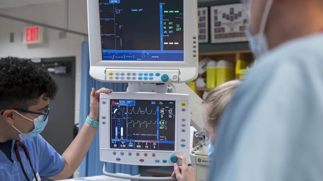 At the School of Health Sciences of Fanshawe College at the London Campus, two people in scrubs and masks are operating a medical ventilator, which has two screens displaying various readings and settings.