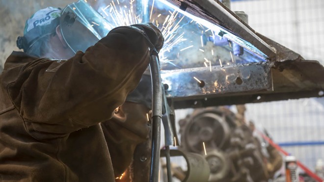 A person wearing protective gear welds metal, creating bright sparks.