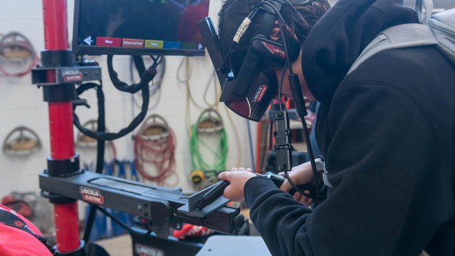 A person uses a virtual reality welding simulator at Fanshawe College, with a Lincoln Electric monitor displaying a welding simulation.