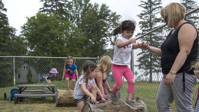 Early Childhood Education program participants are seen on the Simcoe/Norfolk Regional Campus, engaging in outdoor play on a log obstacle course, with a chain link fence and trees in the background.