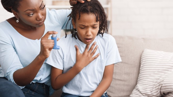 a child sits on a couch struggling to breathe next to his mother who is holding his asthma inhaler prepared to support him.