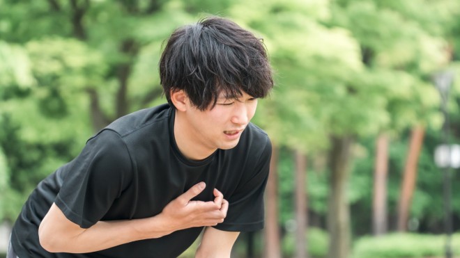 A young male experiences breathing difficulty while on a run outdoors. He appears bent over with a hand on his chest attempting to catch his breathe with blurred trees behind him.