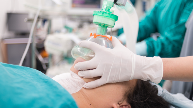 A patent on an operating room table, with a breathing mask over their face being hold in place by an RT