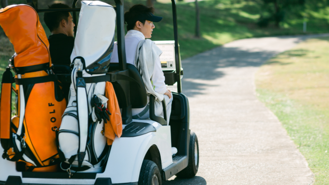 A golf cart on a course pathway, with two golf bags on the back, one orange and one white, two golfers sit in the front